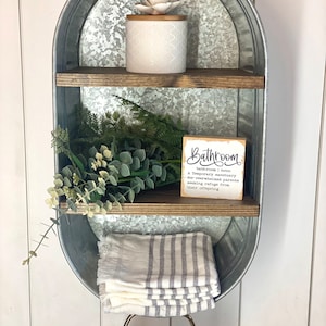 May include: A galvanized metal shelf with two tiers and a towel rack. The shelf is decorated with a small potted plant, a white jar, and a sign that reads "Bathroom | noun | A temporary sanctuary for overwhelmed parents seeking refuge from their offspring."