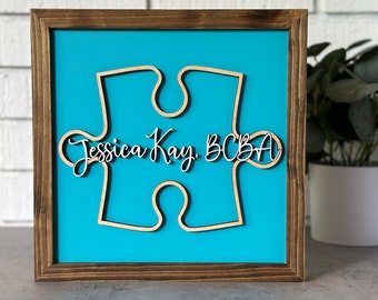 Könnte beinhalten: Ein Holzrahmen mit einem türkisfarbenen Hintergrund und einem ausgeschnittenen Holzpuzzle-Teil. Das Puzzle-Teil trägt den Text "Jessica Kay, BCBA".