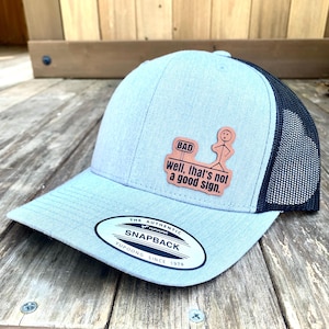 May include: A light blue and black mesh baseball cap with a brown leather patch that says "BAD well, that's not a good sign." The patch features a stick figure standing next to a sign that says "BAD". The cap has a black mesh back and a gray front. The cap has a white label that says "THE AUTHENTIC YUPOONG SNAPBACK YUPOONG SINCE 1974".