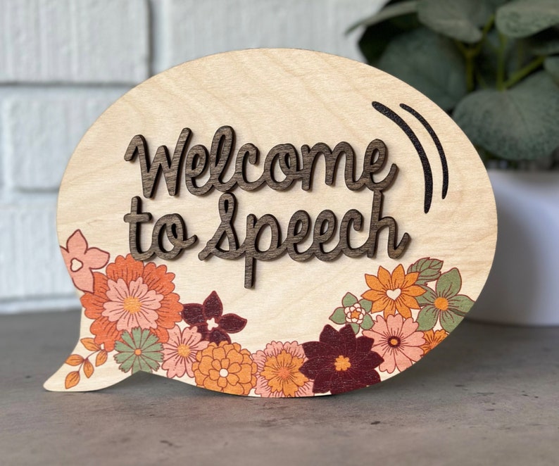 May include: A wooden speech bubble sign with the text "Welcome to Speech" in brown lettering. The sign is decorated with a floral border in shades of orange, pink, and green.