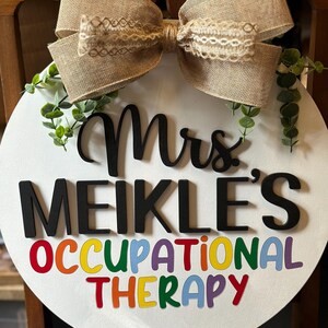 May include: A white round door hanger with a burlap bow and greenery. The text "Mrs. Meikle's Occupational Therapy" is written in black and rainbow letters.