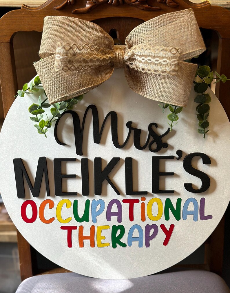 May include: A white round door hanger with a burlap bow and greenery. The text "Mrs. Meikle's Occupational Therapy" is written in black and rainbow letters.