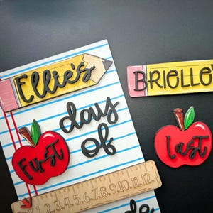 May include: A collection of back-to-school themed wooden cutouts. Includes a pencil with "Ellie's" and "Brielle" on them, a red apple with "First" and "Last", a ruler, and the words "day of".