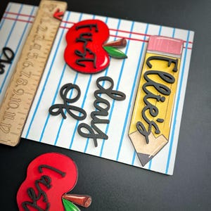 May include: A wooden school-themed decoration with a ruler, two red apples, and a yellow pencil. The apples and pencil have black lettering that reads "First Day" and "Ellie's". The background is a white surface with blue lines.