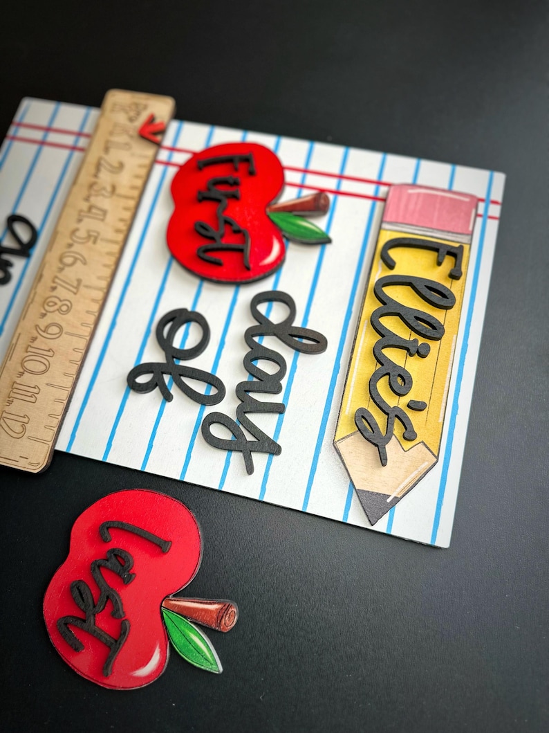 May include: A wooden school-themed decoration with a ruler, two red apples, and a yellow pencil. The apples and pencil have black lettering that reads "First Day" and "Ellie's". The background is a white surface with blue lines.