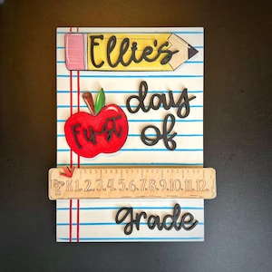 May include: A wooden sign with a school theme. The sign features a pencil with the name "Ellie's", a red apple with "First" on it, and the words "day of grade". A wooden ruler with numbers is also included.