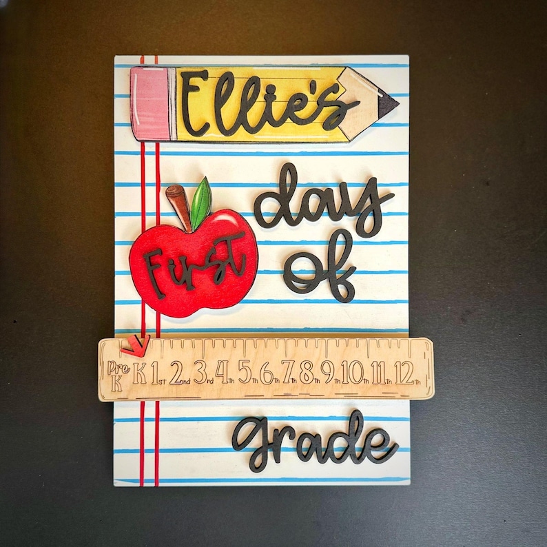 May include: A wooden sign with a school theme. The sign features a pencil with the name "Ellie's", a red apple with "First" on it, and the words "day of grade". A wooden ruler with numbers is also included.