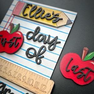 May include: Wooden school-themed decor with a pencil, apples, and ruler. The pencil has the name "Ellie's" in black script. One apple says "first" and the other says "last". The words "day of" are also in black script.