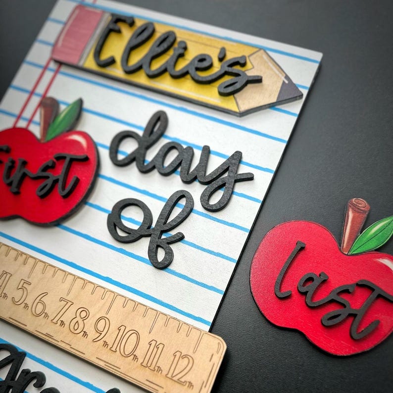 May include: Wooden school-themed decor with a pencil, apples, and ruler. The pencil has the name "Ellie's" in black script. One apple says "first" and the other says "last". The words "day of" are also in black script.