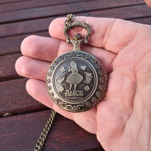 Puede incluir: Un reloj de bolsillo de color bronce con una cadena, con un diseño grabado de Alicia en el País de las Maravillas. La esfera del reloj incluye las palabras "ALICE" y "WONDERLAND" y está rodeada por un patrón floral. El reloj se sostiene en una mano.