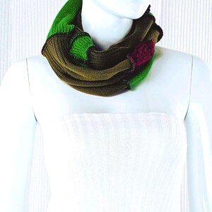 May include: A multi-colored infinity scarf in shades of green, brown, and red. The scarf is made of a soft, knitted material and is draped around a mannequin's neck. The design features a mix of solid and textured sections, creating a visually interesting accessory.