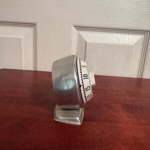 May include: A silver-toned kitchen timer with a rounded metallic casing and a white dial face. The dial features black numerical markings for 5, 10, 15, and 20, with a scalloped edge design. The timer is mounted on a small, square metallic base.