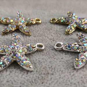Op de afbeelding: Vier zeestervormige bedels met iriserende, veelkleurige strass-steentjes ingelegd op het oppervlak. Twee bedels hebben een goudkleurige metalen basis en twee hebben een zilverkleurige metalen basis. Elke bedel bevat twee kleine lusjes aan de zijkanten.