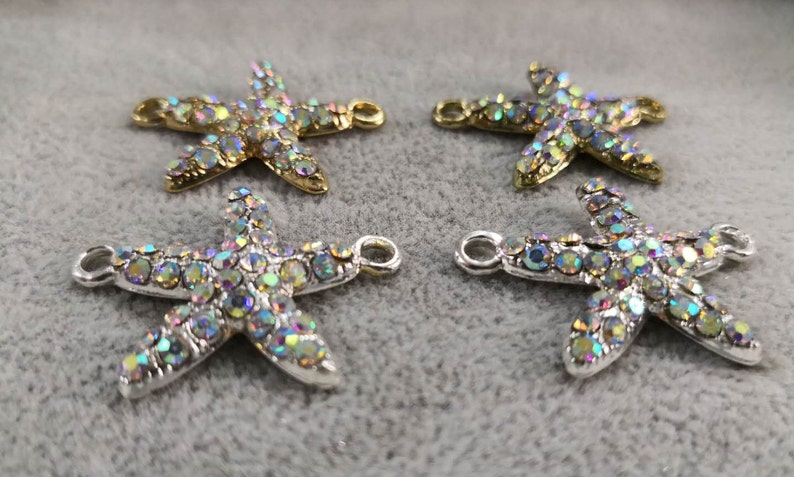 Op de afbeelding: Vier zeestervormige bedels met iriserende, veelkleurige strass-steentjes ingelegd op het oppervlak. Twee bedels hebben een goudkleurige metalen basis en twee hebben een zilverkleurige metalen basis. Elke bedel bevat twee kleine lusjes aan de zijkanten.