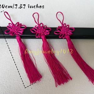 Pode incluir: Três borlas decorativas em fúcsia brilhante com um nó chinês intrincado no topo e um laço para pendurar. O comprimento total de cada borla é de 20 cm, conforme indicado pelo texto e pela linha de medição tracejada na imagem.