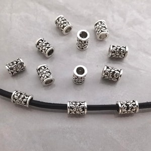 Puede incluir: Cuentas de metal plateadas con un diseño floral. Las cuentas están enhebradas en un cordón de cuero negro.