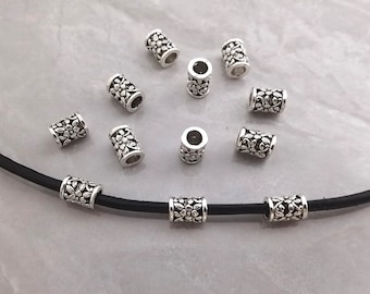 Pode incluir: Uma coleção de contas cilíndricas de metal prateado com um padrão floral em relevo. Oito contas soltas estão dispostas sobre um cordão preto com três contas idênticas enfiadas, exibindo o design detalhado em relevo.