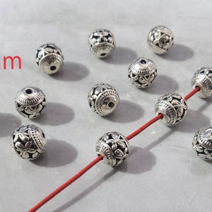 May include: A collection of round, silver-toned metal beads featuring an intricate floral cutout pattern. Each bead measures 10mm in diameter. Several beads are scattered, while three are threaded onto a thin, vibrant red cord for display.