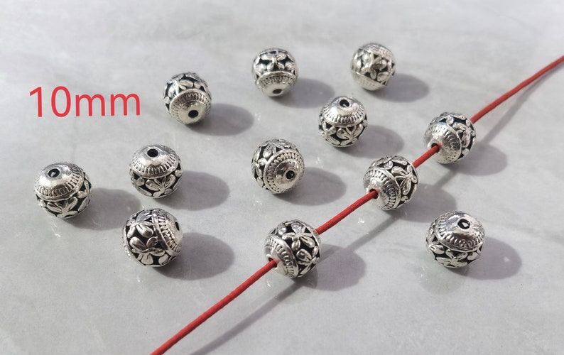 May include: A collection of round, silver-toned metal beads featuring an intricate floral cutout pattern. Each bead measures 10mm in diameter. Several beads are scattered, while three are threaded onto a thin, vibrant red cord for display.