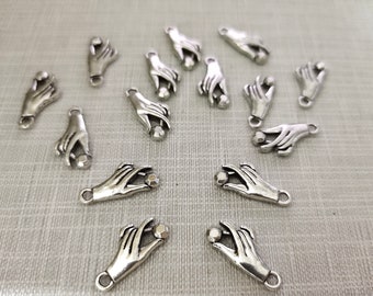 Puede incluir: Una colección de dijes de metal en tono plateado, cada uno con forma de mano sosteniendo una pequeña cuenta esférica. Los dijes cuentan con una anilla en la base para su sujeción y están dispuestos de forma dispersa, mostrando los detalles de los dedos.