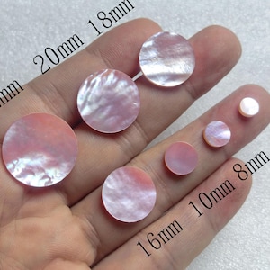 Può includere: Una collezione di dischi iridescenti di madreperla rosa di varie dimensioni, da 6 mm a 22 mm di diametro. I dischi hanno una lucentezza perlacea e sono disposti su una mano.