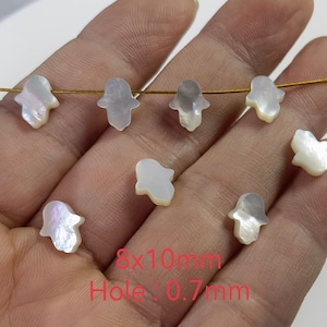 Peut inclure: Huit charmes de main Hamsa en nacre blanche enfilés sur un fil doré. Chaque charme mesure 8 mm sur 10 mm avec un trou de 0,7 mm.