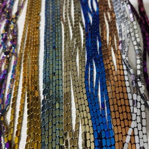 May include: Assortment of colourful, metallic, rectangular beads. The beads are arranged in rows, showcasing a variety of colours, including purple, gold, green, blue, copper, silver, and grey.