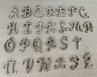 Op de afbeelding: Een collectie van zesentwintig zilverkleurige metalen letterbedels, elk in een cursief lettertype en ingelegd met kleine doorzichtige strass-steentjes. Elke letter van A tot Z is vertegenwoordigd als een individuele hanger met een oogje.