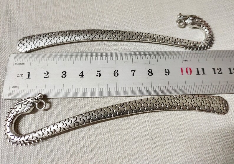 May include: Two silver-toned metal bookmarks featuring a dragon head design with a textured scale pattern along the body. A metal ruler is positioned between the bookmarks, showing a length of approximately 12 centimeters for each item.