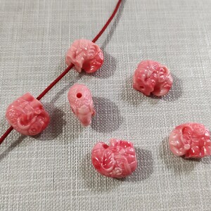 May include: Six pink coral beads carved in the shape of elephants. The beads are smooth and polished, with intricate details.