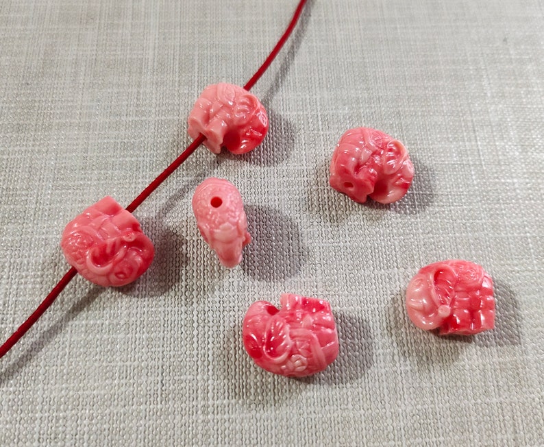 May include: Six pink coral beads carved in the shape of elephants. The beads are smooth and polished, with intricate details.