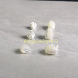 May include: Six white, iridescent, flower-shaped beads arranged in two columns. Each bead features carved petal details with a smooth, glossy finish. The text "Amyjewelry1012" is printed in yellow across the center of the image between the beads.
