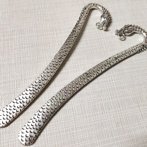 May include: Two silver-toned metal bookmarks featuring a textured scale pattern along the length of the flat blade. Each bookmark terminates in a curved handle shaped like a dragon head, complete with a small circular loop attached to the dragon's snout.