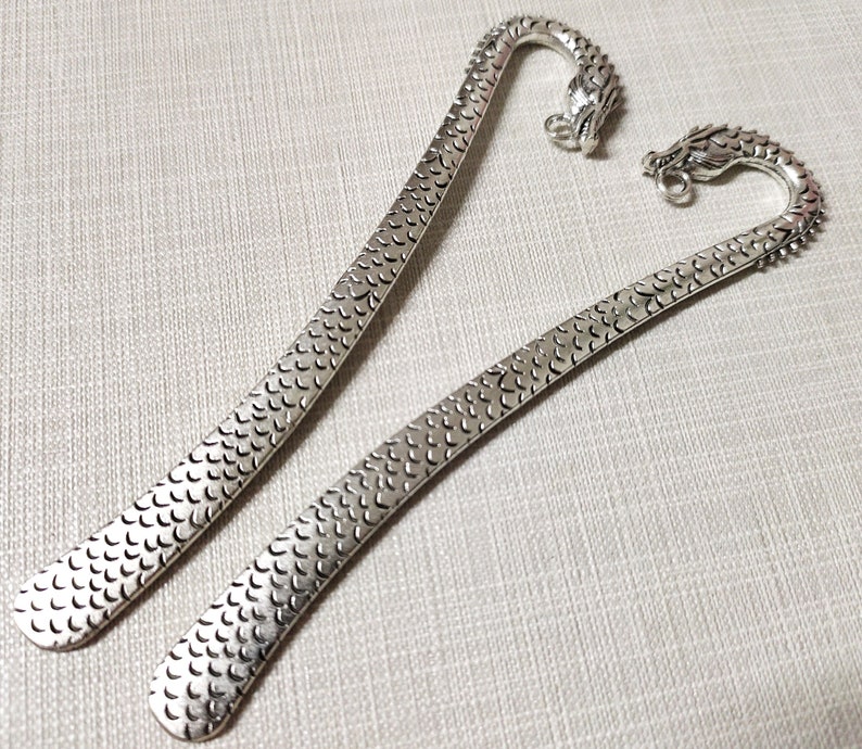 May include: Two silver-toned metal bookmarks featuring a textured scale pattern along the length of the flat blade. Each bookmark terminates in a curved handle shaped like a dragon head, complete with a small circular loop attached to the dragon's snout.