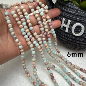 May include: Strands of 6mm gemstone beads in shades of pale blue, beige, and burgundy. The beads are strung together, creating a necklace or bracelet design. The beads have a natural, speckled appearance.