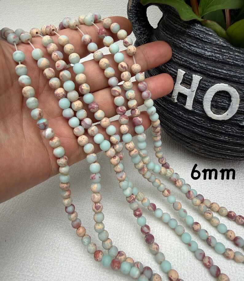 May include: Strands of 6mm gemstone beads in shades of pale blue, beige, and burgundy. The beads are strung together, creating a necklace or bracelet design. The beads have a natural, speckled appearance.