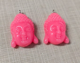 May include: Two pink resin Buddha head pendants with silver jump rings. The pendants are detailed with a serene expression and a textured topknot.