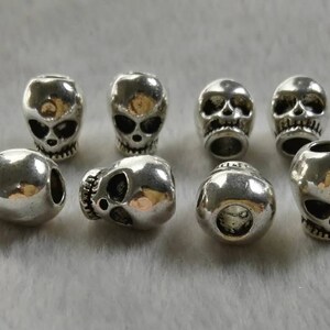 May include: A collection of eight silver-toned metal beads shaped like human skulls. Each bead features detailed eye sockets, a nasal cavity, and teeth. The beads are arranged in two rows, showing various angles including front-facing and side-facing views.