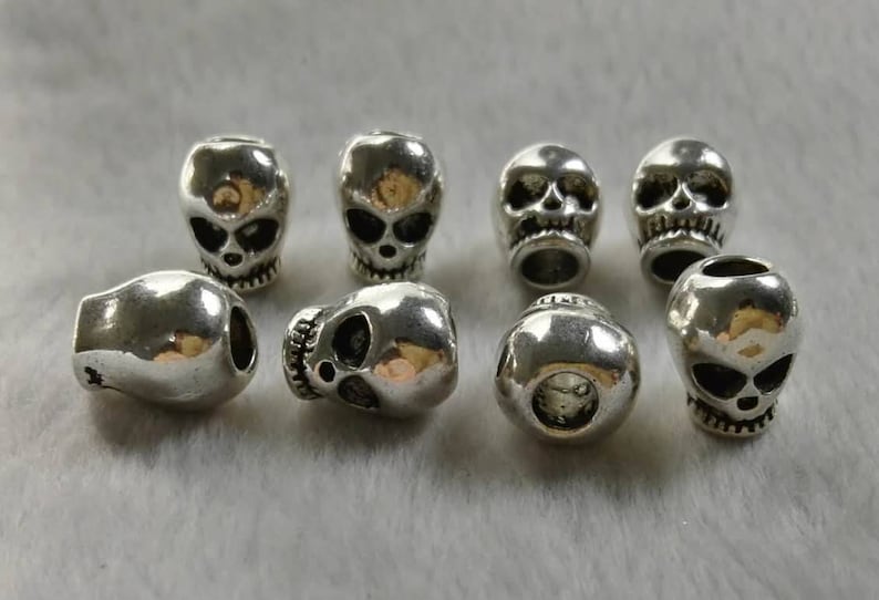 May include: A collection of eight silver-toned metal beads shaped like human skulls. Each bead features detailed eye sockets, a nasal cavity, and teeth. The beads are arranged in two rows, showing various angles including front-facing and side-facing views.