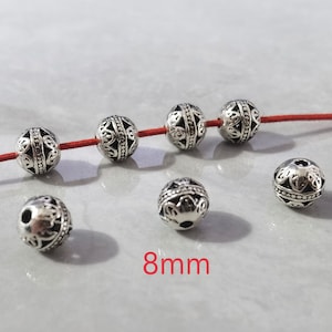 May include: Six round silver-toned metal beads with intricate cut-out filigree patterns and a central horizontal band. Four beads are threaded on a thin red cord, while two are loose. The text 8mm is printed in red at the bottom center.