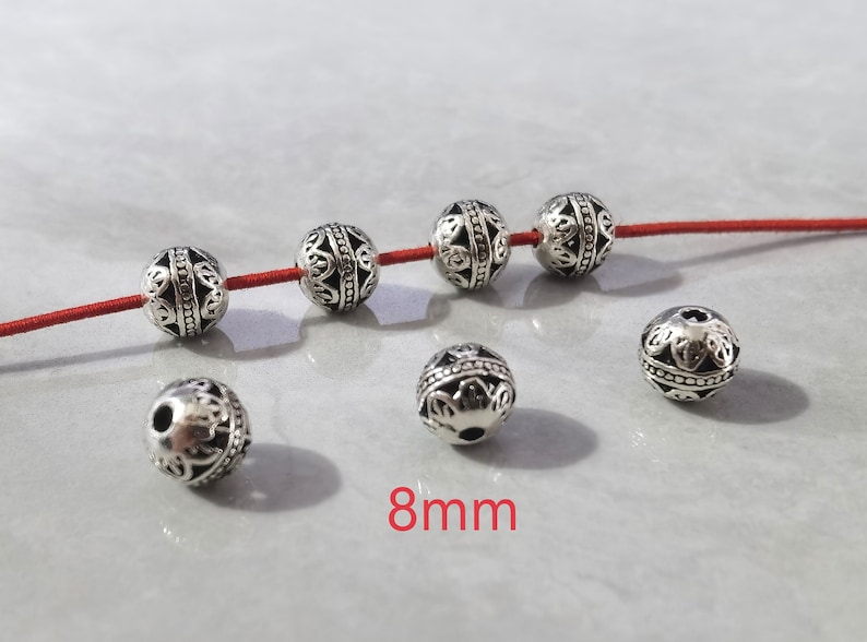 May include: Six round silver-toned metal beads with intricate cut-out filigree patterns and a central horizontal band. Four beads are threaded on a thin red cord, while two are loose. The text 8mm is printed in red at the bottom center.