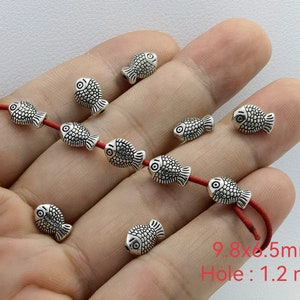 Könnte beinhalten: Silberne fischförmige Perlen mit einem roten Faden, der durch sie gefädelt ist. Die Perlen sind etwa 9,8 mm x 6,5 mm groß und haben ein Loch von 1,2 mm.