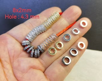 Op de afbeelding: Een collectie ronde, platte tussenkralen ingelegd met heldere zirkonia-steentjes in goud-, zilver-, roségoud- en gunmetal-tinten. De kralen meten 8x2mm met een gat van 4,3 mm. Sommige kralen zijn geregen aan een rood koord.