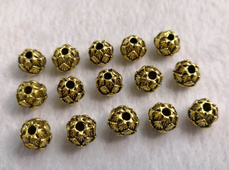 May include: A collection of fourteen small, round, gold-toned metal beads. Each bead features a detailed, textured surface with a repeating petal-like pattern and a central hole for threading, arranged in a loose group on a white surface.
