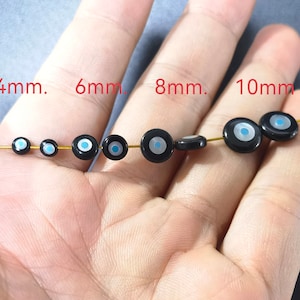 May include: A close-up of a hand holding a string of black beads with a blue and white evil eye design. The beads are in different sizes, ranging from 4mm to 10mm. The text "4mm. 6mm. 8mm. 10mm" is printed above the beads.