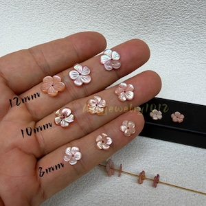 May include: A collection of light pink, iridescent, carved shell flower beads arranged by size. The beads are labeled 12mm, 10mm, and 8mm, indicating their respective diameters. Each flower-shaped bead features a central hole for stringing.