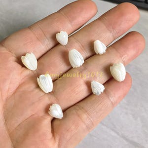 May include: Eight small, white, carved shell beads shaped like tulip flower buds. Each bead features vertical ridges and a smooth, iridescent finish. The beads are arranged in a scattered pattern, showcasing their delicate, organic form and pearlescent luster.