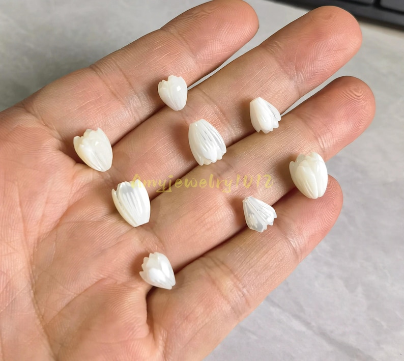 May include: Eight small, white, carved shell beads shaped like tulip flower buds. Each bead features vertical ridges and a smooth, iridescent finish. The beads are arranged in a scattered pattern, showcasing their delicate, organic form and pearlescent luster.