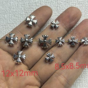 Op de afbeelding: Een collectie zilverkleurige metalen kruiskralen met een decoratief reliëfontwerp. De kralen zijn verkrijgbaar in twee maten, waarbij sommige 12x12mm meten en andere 8,5x8,5mm, zoals aangegeven door de rode tekst op de afbeelding.