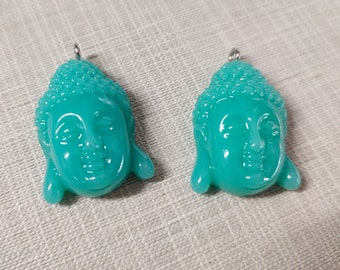 May include: Two turquoise Buddha head pendants with a silver loop for hanging. The pendants have detailed facial features and textured hair. The pendants are made of a glossy material.